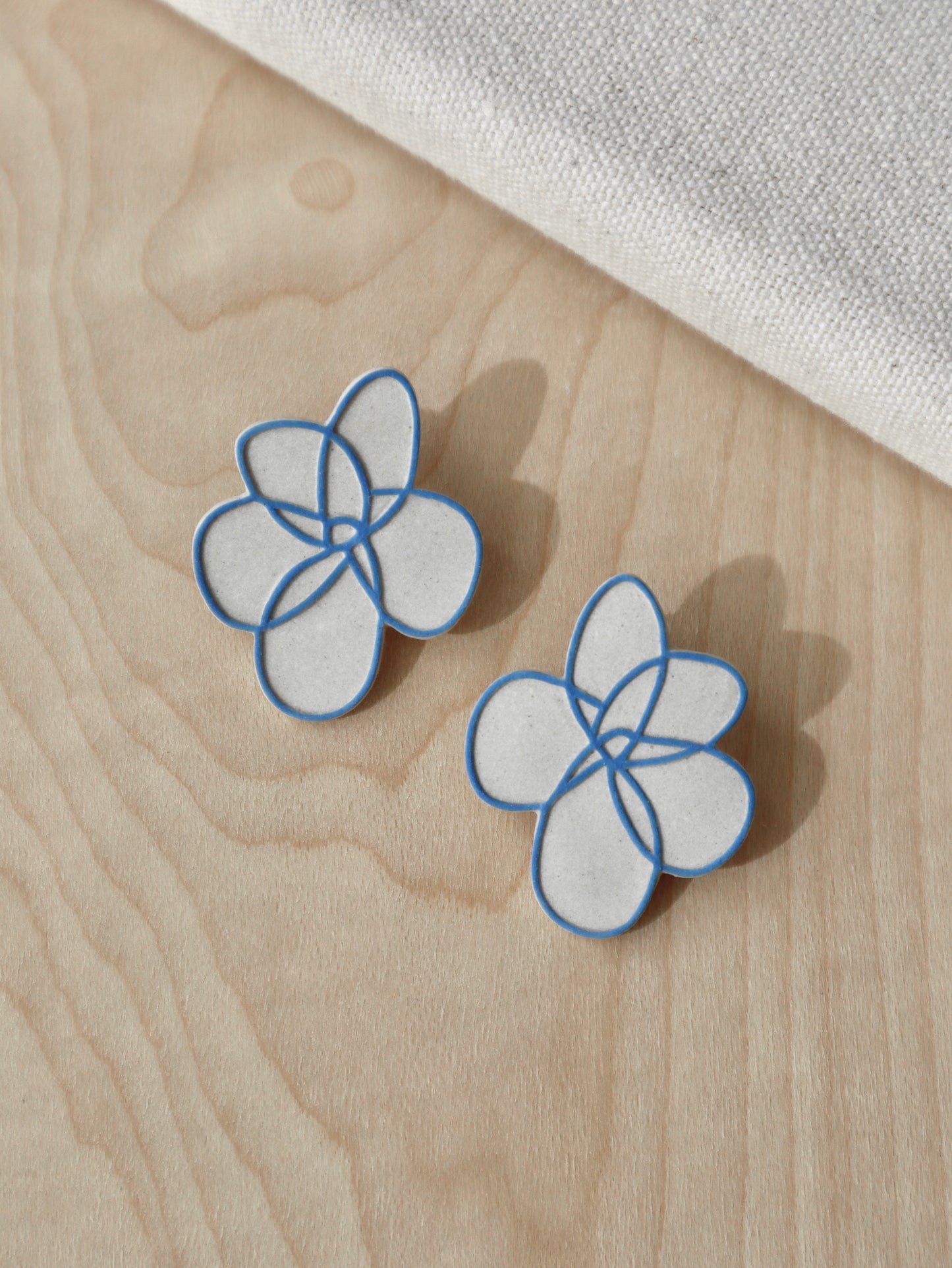 Handcrafted beige stoneware flower stud earrings with painted blue outline design.