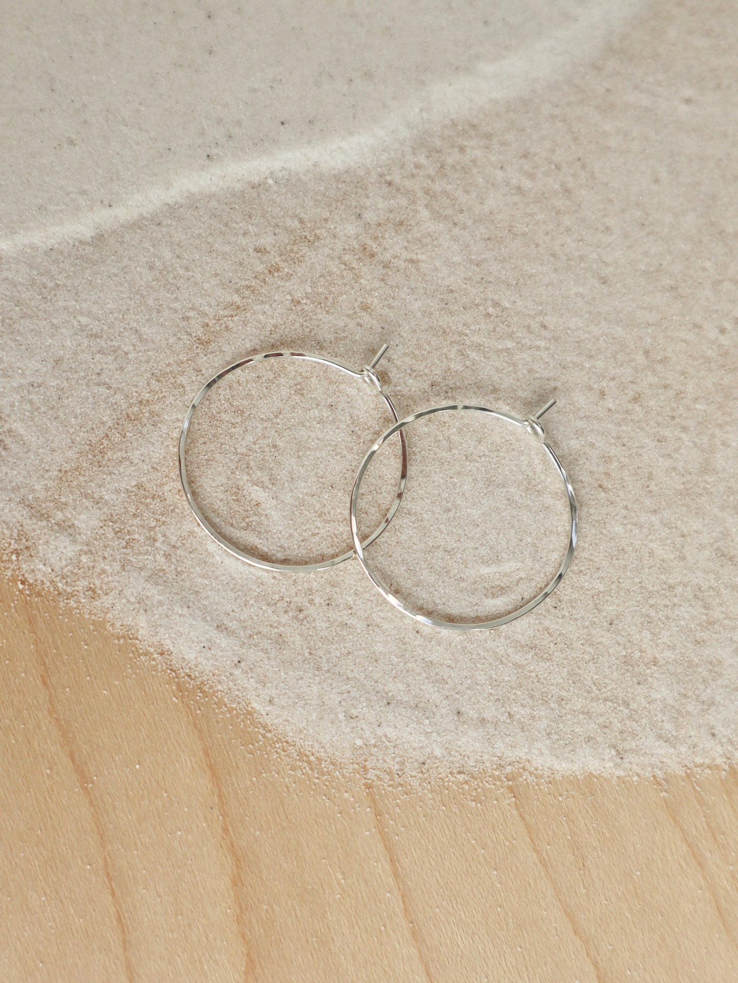 A pair of thin sterling silver hoop earrings with a hand-hammered texture that catches the light. The ear wire tucks into a small loop, holding it in place.