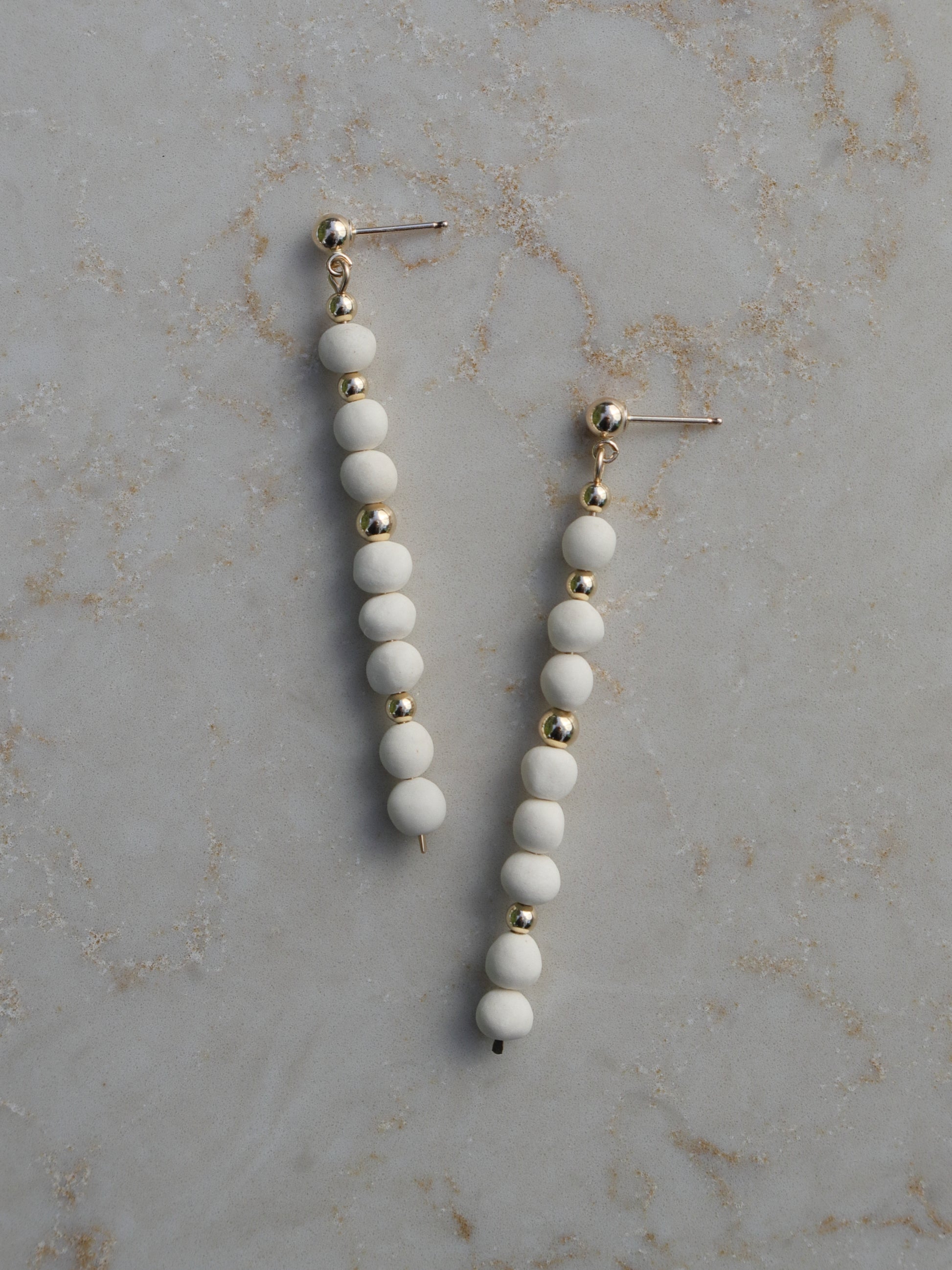 Long, dangle earrings made with stacked white porcelain and gold beads hanging on ball posts.